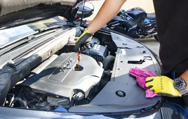 A person wearing yellow work gloves pulls a dipstick to check the engine oil level in a modern car engine bay.