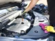 A person wearing yellow work gloves pulls a dipstick to check the engine oil level in a modern car engine bay.