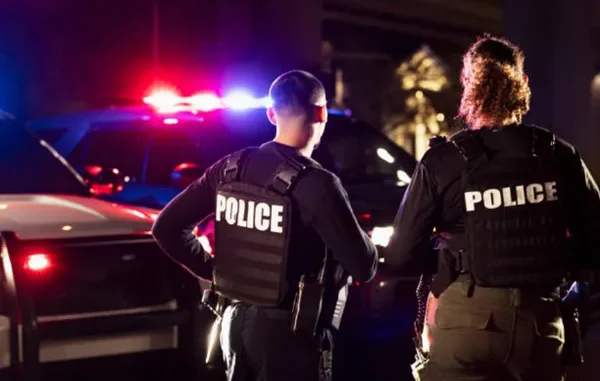 Two police officers in tactical vests standing in front of a patrol car with flashing red and blue lights at night.