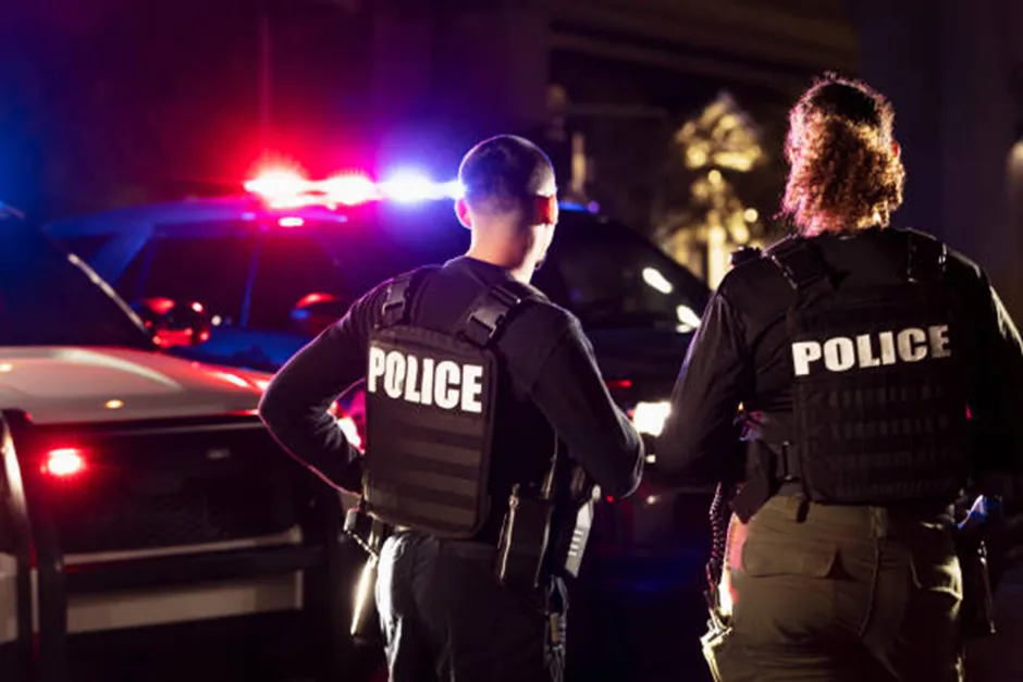 Two police officers in tactical vests with "POLICE" written on the back stand in front of patrol cars with flashing red and blue lights at night.