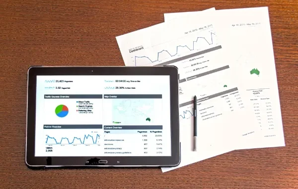 A tablet displaying a financial dashboard with pie charts and line graphs next to printed data reports and a stylus on a wooden desk.