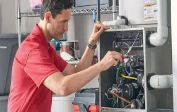 A professional HVAC technician in a red shirt inspecting the internal wiring and components of a residential furnace.