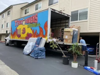 A Trek Movers truck parked at an apartment complex with its liftgate open, showing boxes, furniture, and a person carefully loading items.