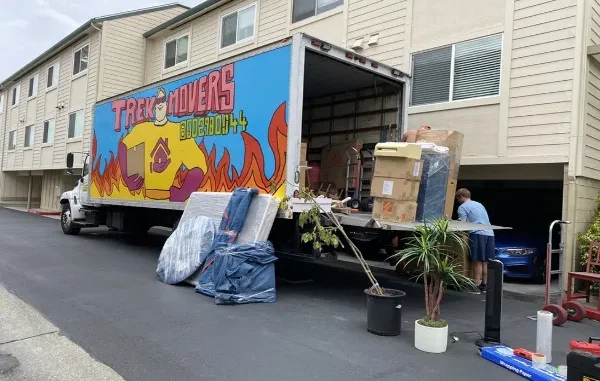 A Trek Movers truck parked at an apartment complex with its liftgate open, showing boxes, furniture, and a person carefully loading items.