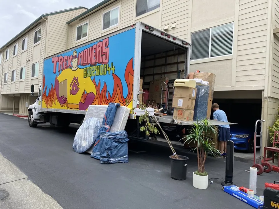 A professional Trek Movers box truck parked outside an apartment building with movers loading furniture, boxes, and potted plants onto a lift gate.