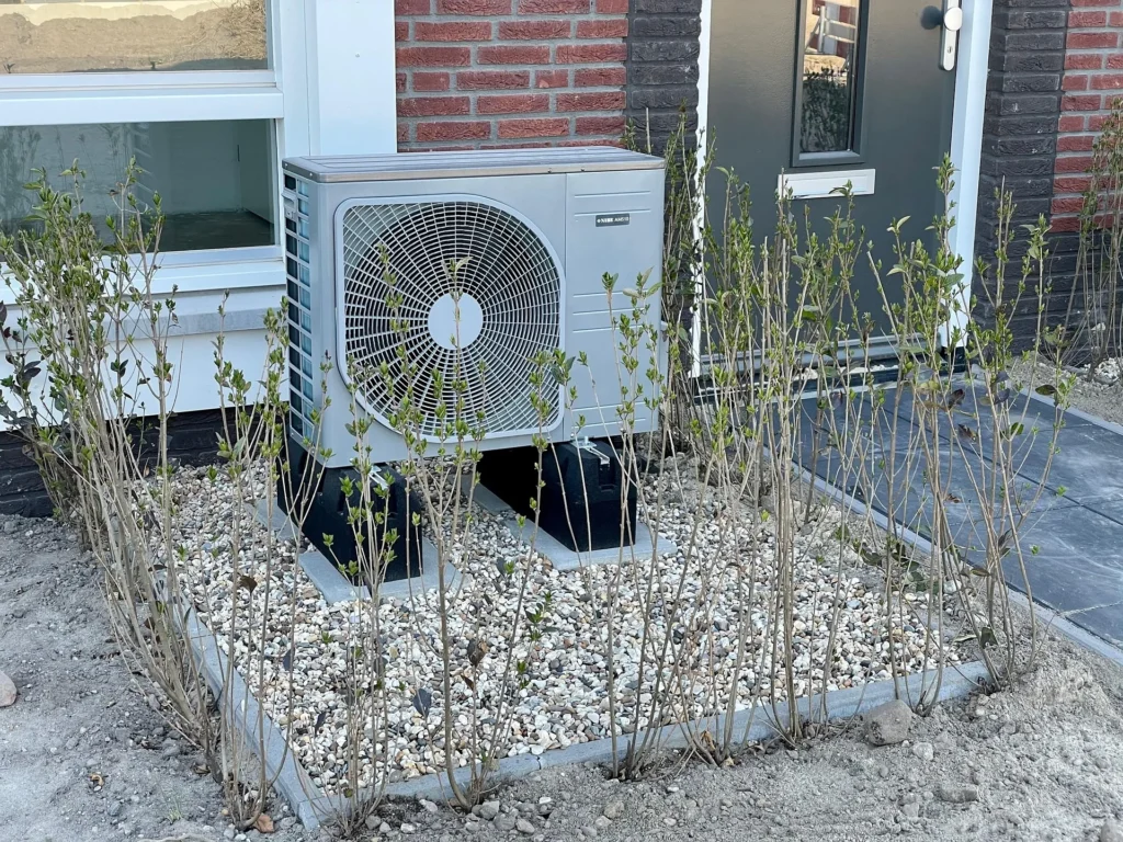 An outdoor air-source heat pump unit installed on elevated vibration-damping mounting blocks atop a gravel bed, positioned with proper clearance from the home exterior to ensure optimal airflow and long-term performance.