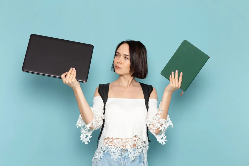 A young professional woman thoughtfully comparing a sleek black laptop in one hand and a green notebook in the other against a light blue background, symbolizing the choice between traditional and digital professional tools.