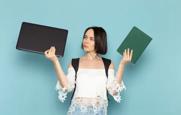 A young professional woman thoughtfully comparing a sleek black HP laptop in one hand and a green hardcover notebook in the other against a light blue background.