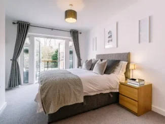 A clean, minimalist bedroom featuring a well-made bed with grey linens and open French doors leading to a balcony, letting in fresh air.