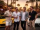 Four stylishly dressed men holding drinks and laughing in front of luxury sports cars on a palm-tree-lined street in Scottsdale at sunset.