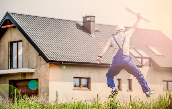 A construction worker in blue overalls and a hard hat jumping for joy in a field, holding rolled blueprints, with a renovated modern house in the background.