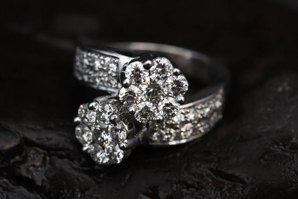 A luxurious "Toi et Moi" style diamond ring featuring two large flower-shaped diamond clusters on a pavé-set white gold band, displayed against a dark, textured background.