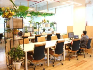 A bright, modern shared office space with long white desks, ergonomic tan leather chairs, and black metal shelving units filled with green potted plants.