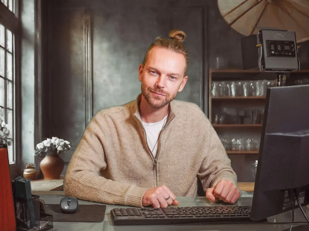 A professional man with a man-bun and beige sweater sitting at a computer desk with a keyboard and monitor, smiling confidently in a modern home office setting.