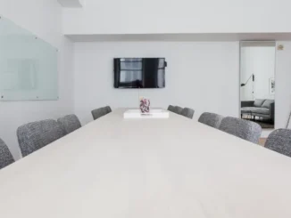A long, light wood conference table surrounded by grey chairs in a clean, white modern office with a TV and glass whiteboards.