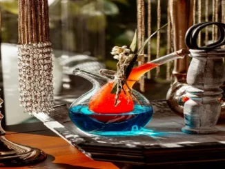 A close-up of a decorative, hand-blown glass shisha base featuring a dual-chamber design with vibrant blue and orange liquids. The setup is adorned with dried flowers and twine, set against a backdrop of golden bead curtains and rustic wooden decor in a sunlit Dubai lounge.