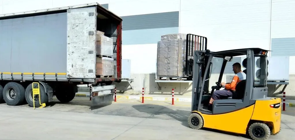A yellow forklift operator loading large palletized goods into the back of a freight truck at a warehouse loading dock. This demonstrates the logistics and fulfillment stage of the final mile delivery process.
