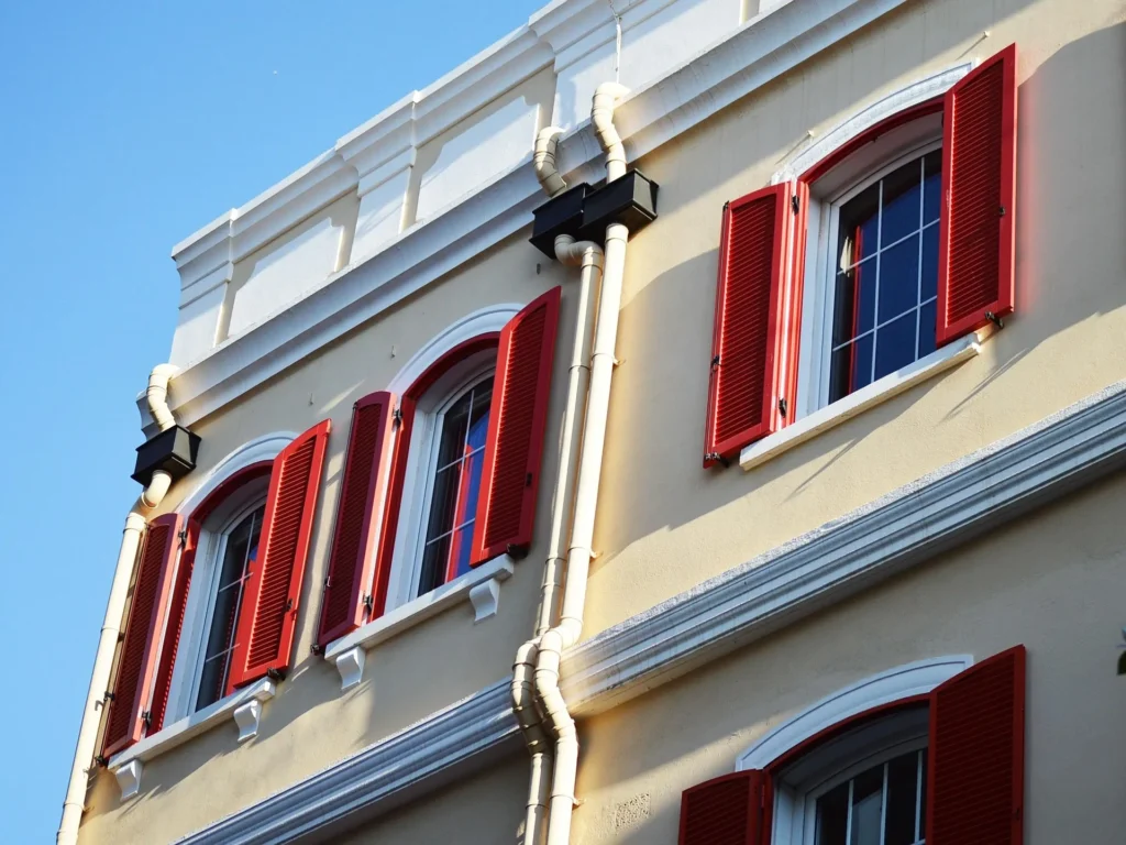 A close-up, low-angle shot of a tan building exterior featuring two vertical white downspouts neatly secured to the wall. The downspouts are positioned between arched windows with bright red shutters, demonstrating a clean and functional drainage system designed to direct rainwater away from the building’s foundation.