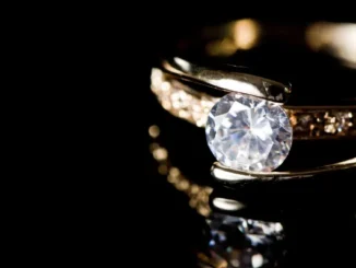 A close-up shot of a brilliant-cut diamond engagement ring with a gold band and pave-set side stones, reflecting on a polished black surface.