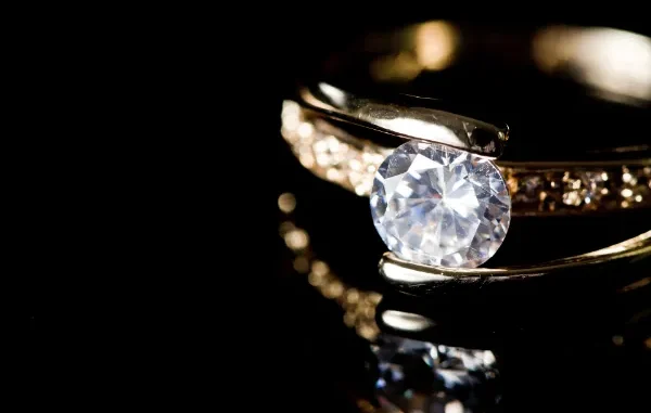 A close-up shot of a brilliant-cut diamond engagement ring with a gold band and pave-set side stones, reflecting on a polished black surface.