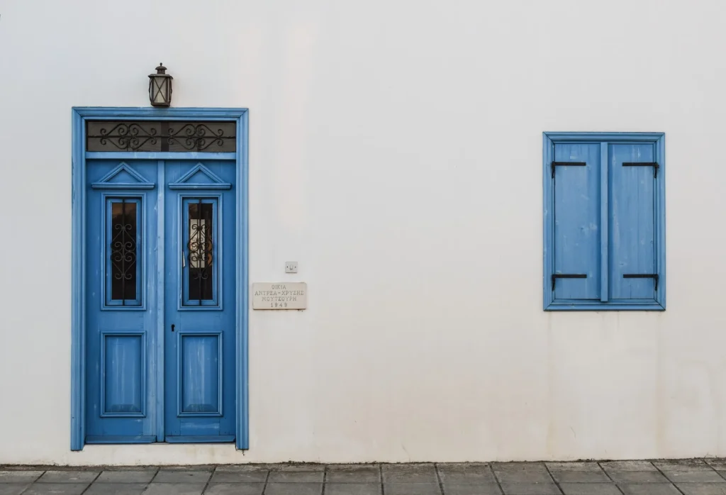 A minimalist white stucco exterior of a Mediterranean-style building featuring a vibrant blue wooden door and matching shutters. The roofline lacks a gutter system, illustrating a scenario where rainwater can fall directly onto the base of the structure, potentially leading to foundation erosion.