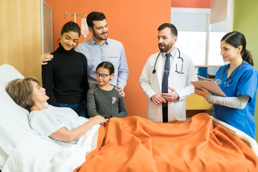 A woman that had a stroke lies in a hospital bed surrounded by her family while a male doctor and female nurse discuss her care.