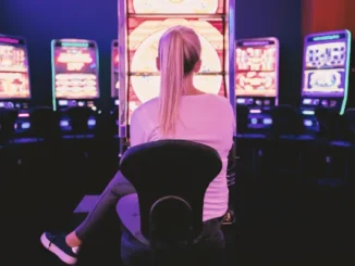 A woman with blonde hair tied in a ponytail sits in front of a brightly lit, neon slot machine in a dark casino, focused on the glowing game screen.
