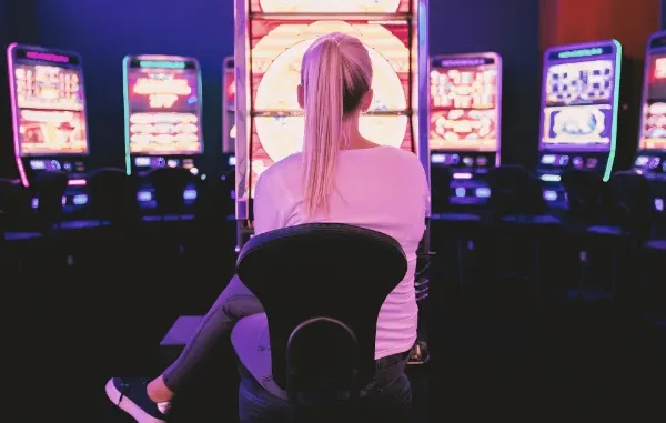 A woman with blonde hair tied in a ponytail sits in front of a brightly lit, neon slot machine in a dark casino, focused on the glowing game screen.