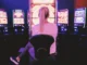 A woman with blonde hair tied in a ponytail sits in front of a brightly lit, neon slot machine in a dark casino, focused on the glowing game screen.
