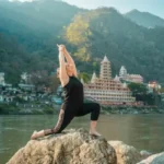 A woman practicing a low lunge yoga pose on a rock by the Ganges River in Rishikesh, with the iconic multi-story temples and Himalayan foothills in the background.