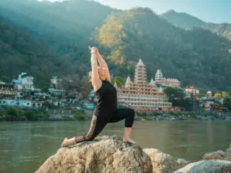 A woman practicing a low lunge yoga pose on a rock by the Ganges River in Rishikesh, with the iconic multi-story temples and Himalayan foothills in the background.