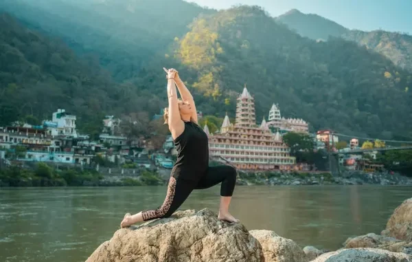 A woman practicing a low lunge yoga pose on a rock by the Ganges River in Rishikesh, with the iconic multi-story temples and Himalayan foothills in the background.