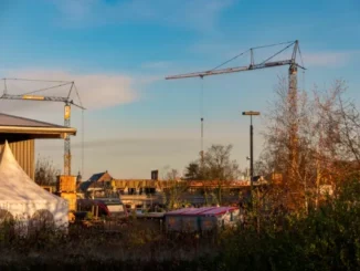 A construction site at golden hour featuring two large tower cranes, a white event tent, and a residential building under construction against a clear blue sky.
