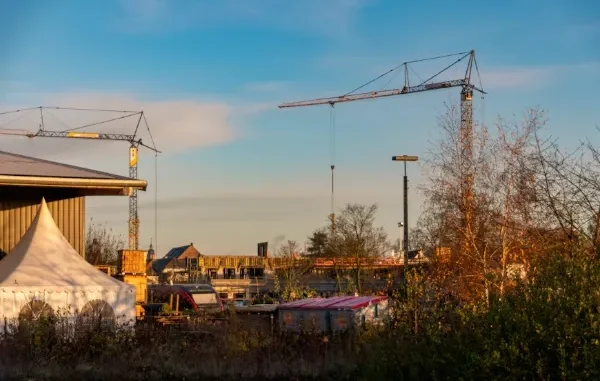 A construction site at golden hour featuring two large tower cranes, a white event tent, and a residential building under construction against a clear blue sky.