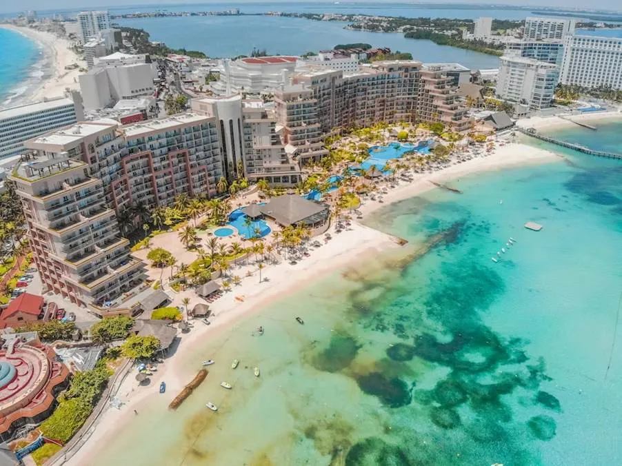 A high-angle view of a luxury Cancun resort featuring turquoise swimming pools, palm trees, and a white sandy beach nestled between the Caribbean Sea and Nichupté Lagoon.
