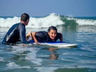 A smiling woman lies on a blue and white surfboard in the ocean while a male instructor in a wetsuit guides her through a breaking wave.