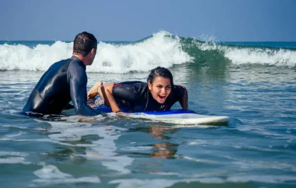 A smiling woman lies on a blue and white surfboard in the ocean while a male instructor in a wetsuit guides her through a breaking wave.