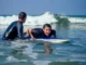 A smiling woman lies on a blue and white surfboard in the ocean while a male instructor in a wetsuit guides her through a breaking wave.