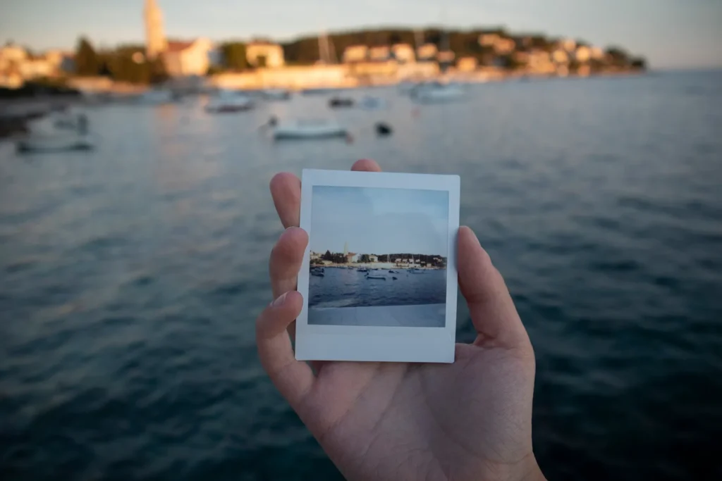 A hand holding a square instant photo that perfectly frames a coastal Mediterranean town with blue water, illustrating subject isolation for AI background removal.