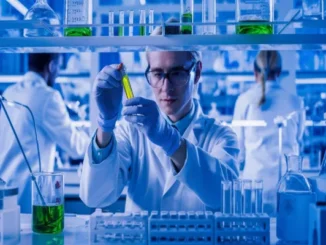 A laboratory technician in a white coat, safety glasses, and blue gloves holds up a test tube containing a vibrant green liquid for close inspection in a high-tech chemical laboratory with blue ambient lighting.