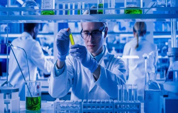 A laboratory technician in a white coat, safety glasses, and blue gloves holds up a test tube containing a vibrant green liquid for close inspection in a high-tech chemical laboratory with blue ambient lighting.
