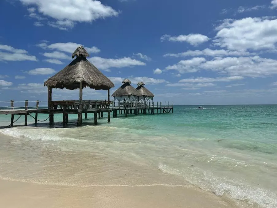 A wooden pier with thatched-roof palapas extending over turquoise Caribbean water on a white sand beach in Cancun.