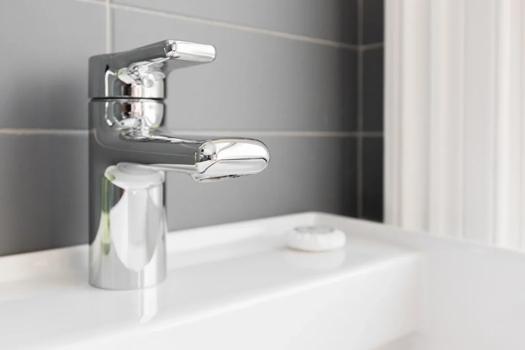 Close-up of a polished chrome faucet on a white ceramic sink in a clean bathroom with grey tiled walls.