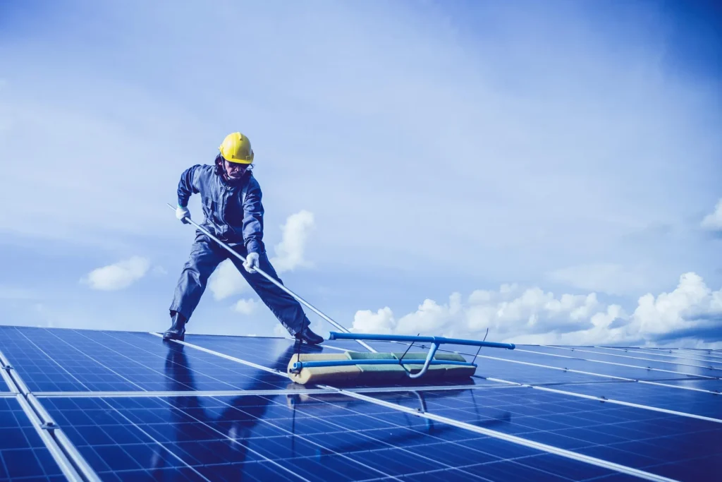 A maintenance worker in a yellow hard hat and protective gear using a specialized long-reach brush to clean a large array of rooftop solar panels under a clear blue sky.