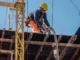 A construction worker wearing a yellow hard hat and a safety harness secured to a lifeline while working on an elevated concrete formwork platform.