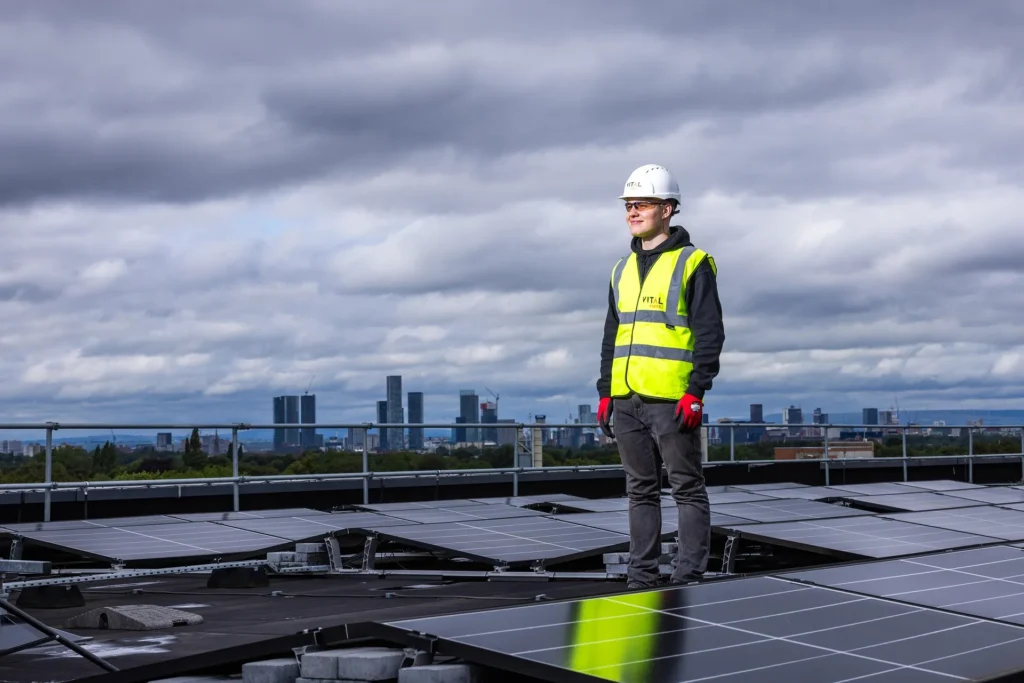 A safety professional wearing a white hard hat, high-visibility vest, and protective gloves stands on a commercial rooftop amidst a large solar panel array, with a city skyline in the background.
