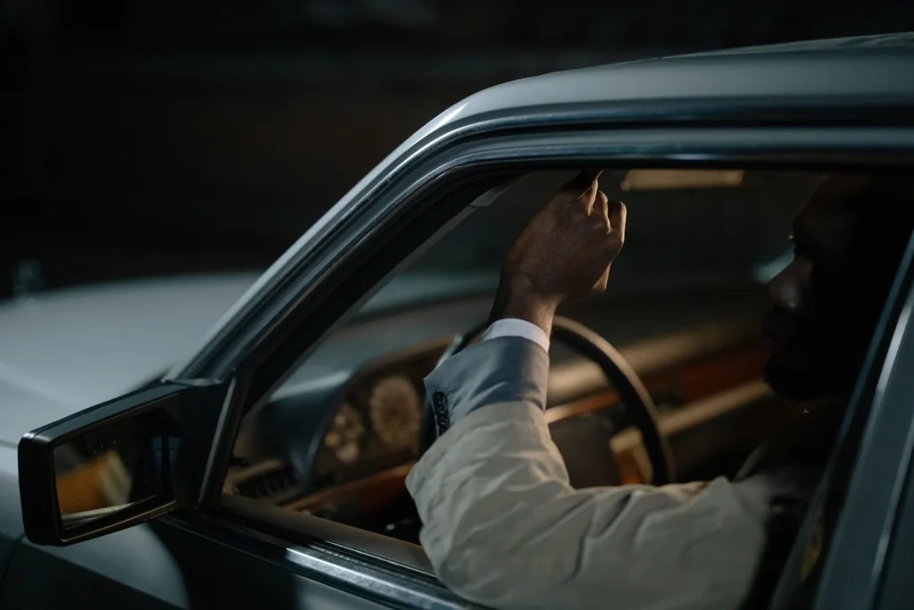 A driver in a business suit adjusting the rearview mirror of a car at night, demonstrating proper arm reach and ergonomic positioning to reduce back pain during travel.