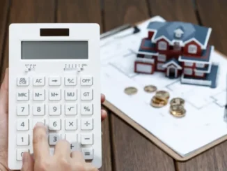 A person's hand using a white calculator next to a small model house, gold coins, and a house blueprint on a clipboard.