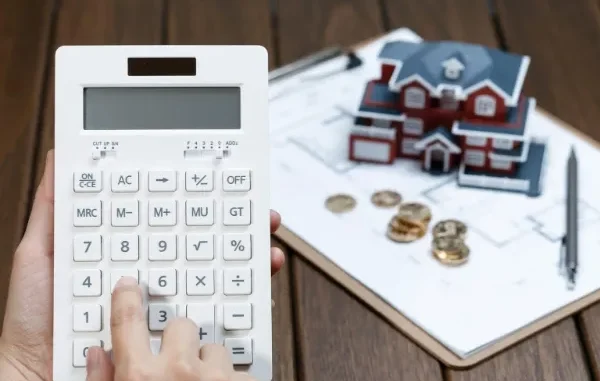 A person's hand using a white calculator next to a small model house, gold coins, and a house blueprint on a clipboard.