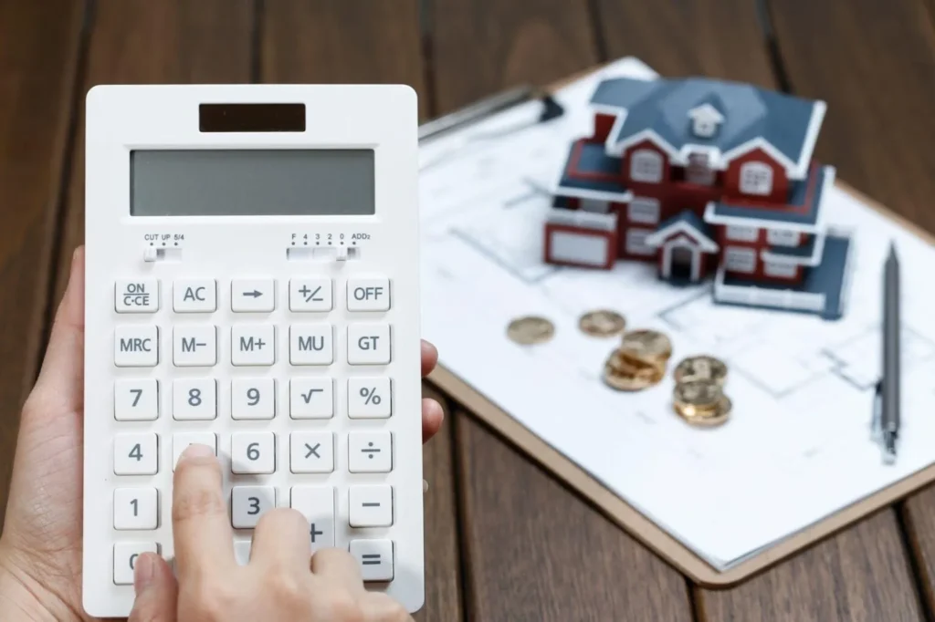 A person's hand using a white calculator next to a small house model and gold coins on a clipboard with blueprints.